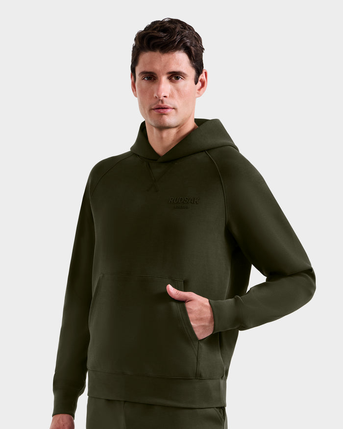 A man stands facing forward with one hand in the pocket of a Rudsak BARRY - 9524011 Dark Olive M oversized hoodie, featuring a front pocket, against a plain light background.