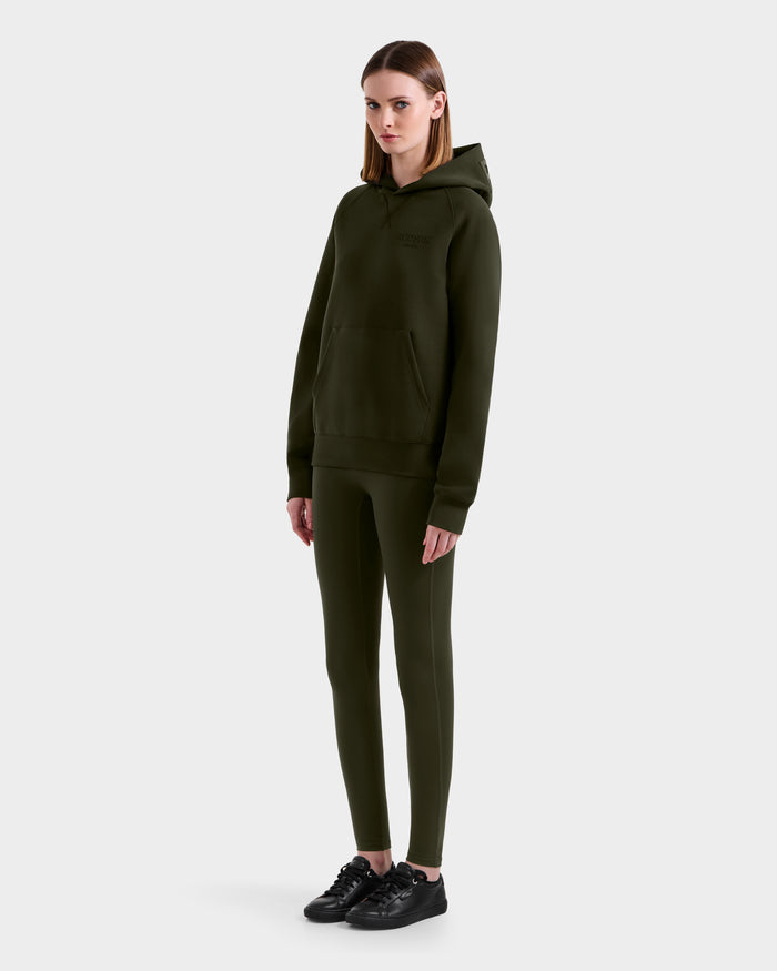 A woman with straight, shoulder-length hair and a neutral expression wears the Rudsak BARRY - 9524011 Dark Olive W oversized hoodie and matching leggings, paired with black sneakers, standing against a plain background with arms relaxed at her sides.