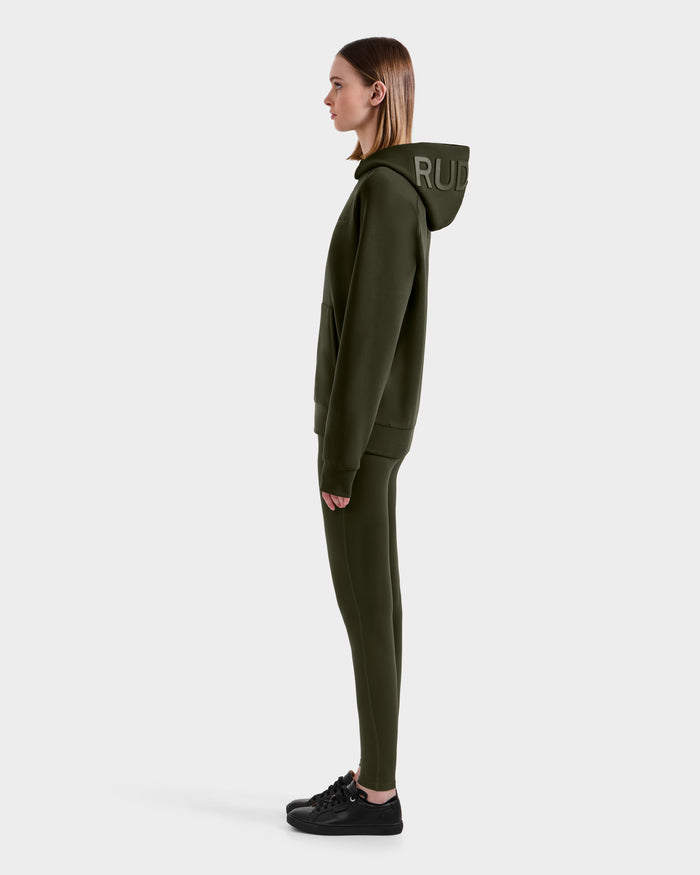 A woman in profile wears a Rudsak BARRY - 9524011 Dark Olive W oversized hoodie with the hood up, matching leggings, and black sneakers, standing against a plain white background.