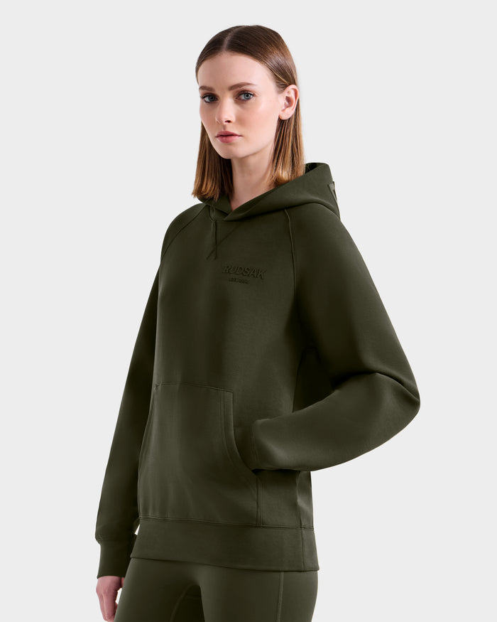 A woman with straight, shoulder-length brown hair wears the Rudsak BARRY - 9524011 hoodie and matching pants in dark olive, standing against a plain light gray background.