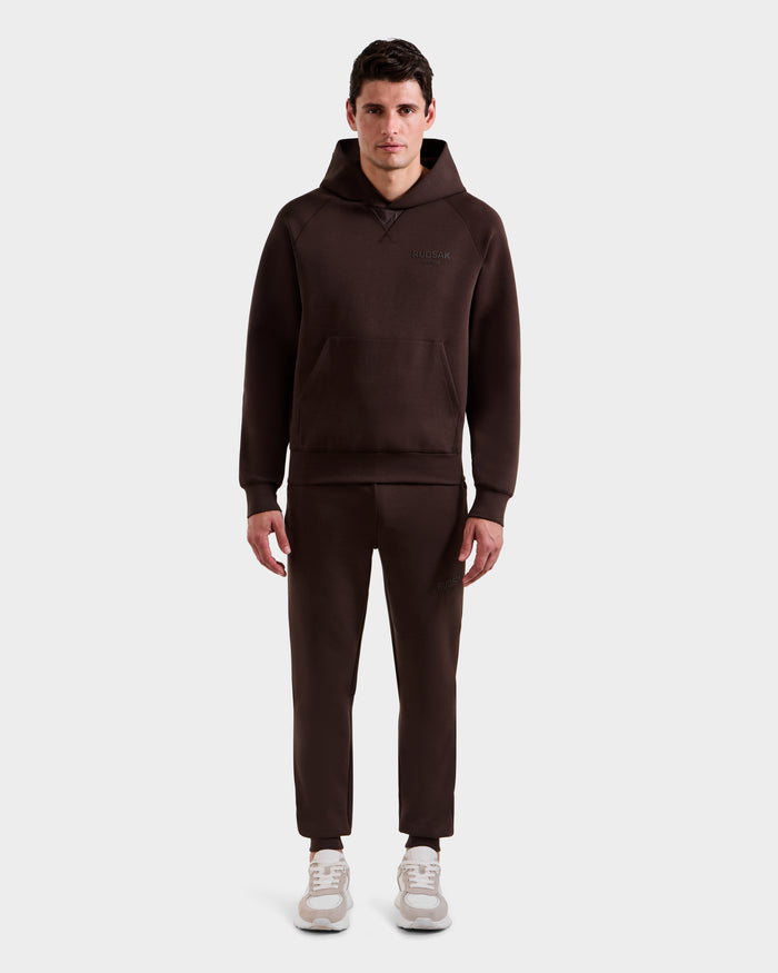 A man stands facing forward against a white background, wearing the Rudsak BARRY - 9524011 Espresso M hoodie and matching jogger pants, paired with light-colored sneakers.
