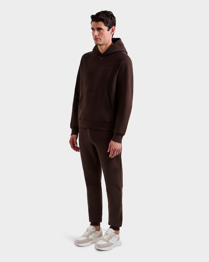 A man stands against a simple light background wearing the Rudsak BARRY - 9524011 Espresso M hoodie and matching jogger pants, paired with white and beige sneakers. He faces slightly to the side with a neutral expression.