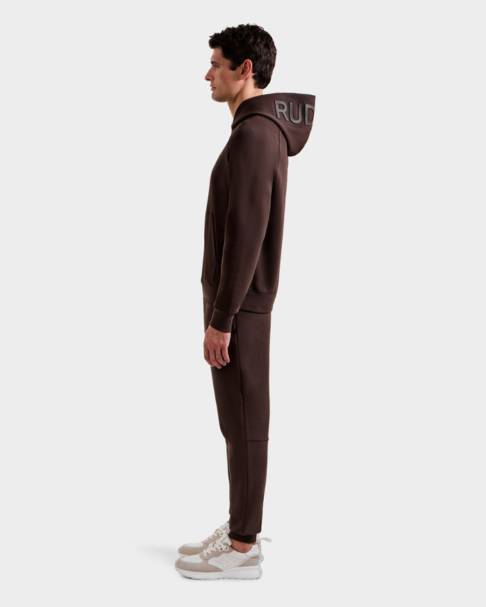 A man stands in profile wearing the Rudsak BARRY - 9524011 Espresso M unisex hoodie with "RUD" on the hood, matching brown joggers, and white sneakers against a plain light background.