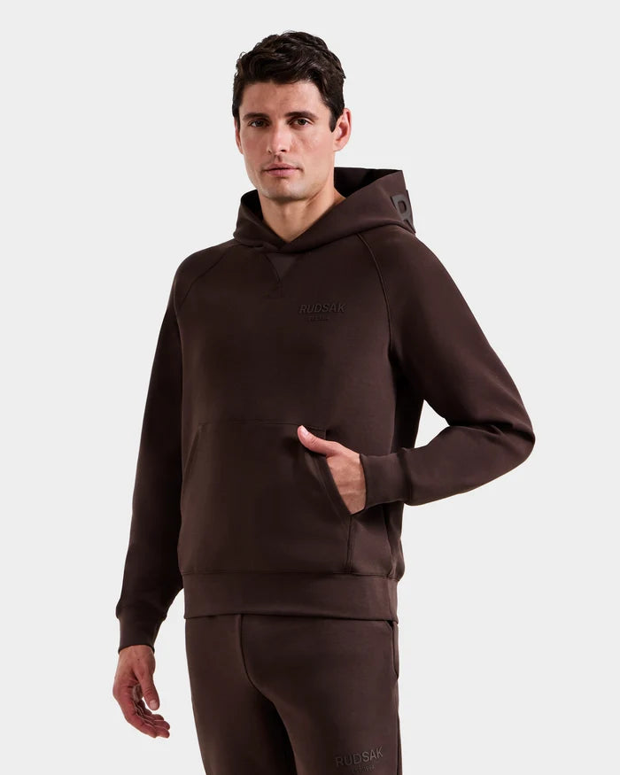 A man wears the BARRY - 9524011 Espresso M set by Rudsak, featuring a dark brown unisex hoodie with subtle branding and matching sweatpants, standing against a plain white background with one hand in his pocket.