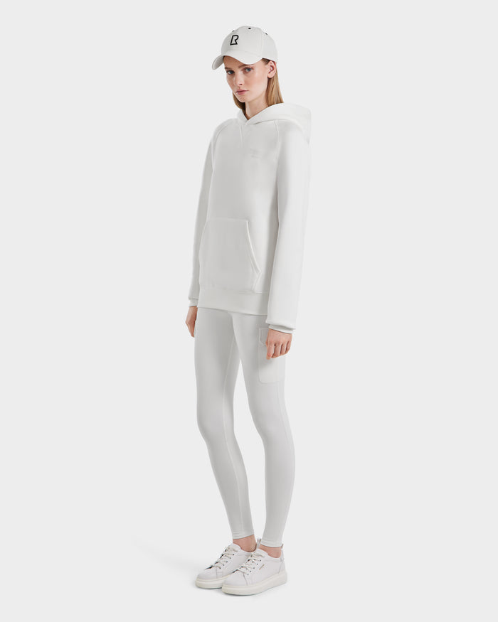 A person stands against a white background wearing the Rudsak BARRY - 9524011 Iceland W oversized hoodie, paired with white leggings, a white cap, and white sneakers. Their expression is neutral and their hands are relaxed by their sides.