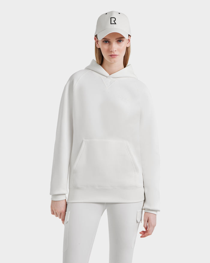 A woman in an all-white outfit—oversized unisex Rudsak BARRY - 9524011 Iceland W hoodie, matching pants, and a white baseball cap with a black R logo—stands against a plain white background.