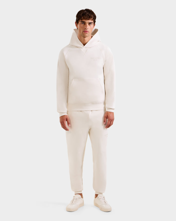 A man faces forward wearing a Rudsak BARRY - 9524011 Vanilla M unisex hoodie and matching jogger pants with off-white sneakers, set against a plain light background.