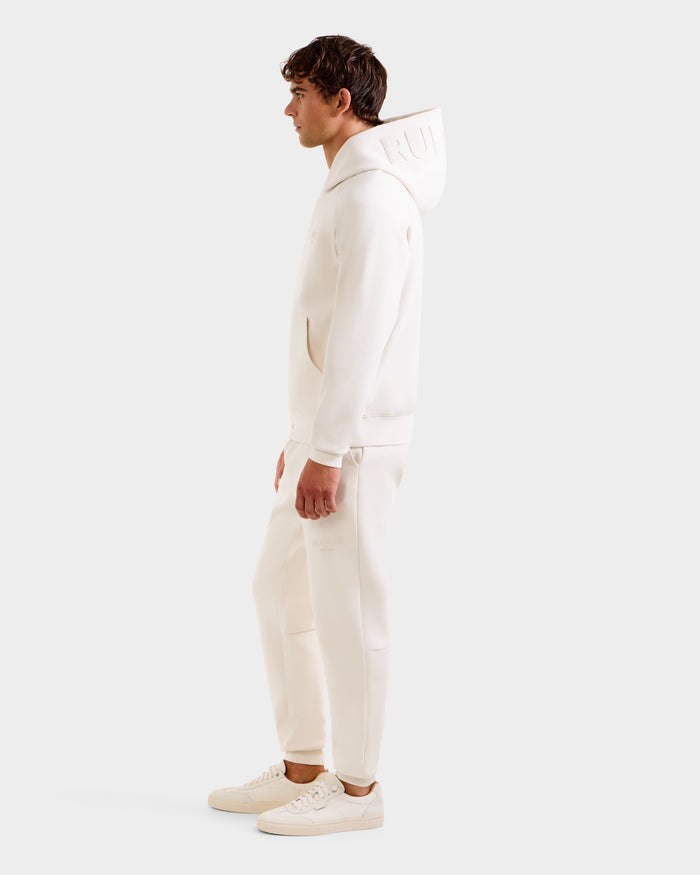 A man stands in profile wearing the Rudsak BARRY - 9524011 Vanilla M unisex hoodie and matching joggers with off-white sneakers against a plain light background.