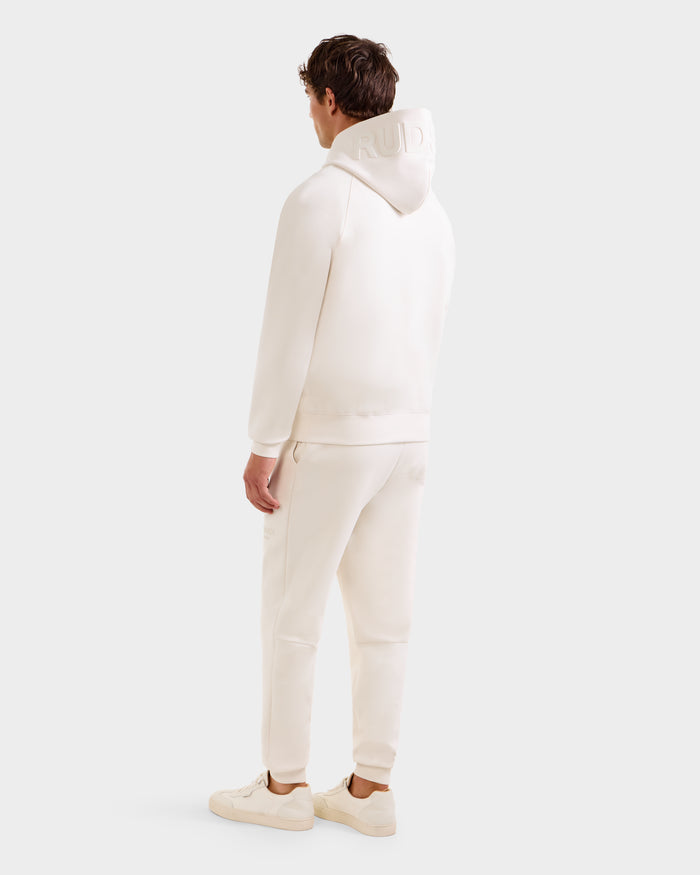A man faces away, wearing the Rudsak BARRY - 9524011 Vanilla M unisex hoodie with matching joggers and sneakers, hood up, against a plain light background.