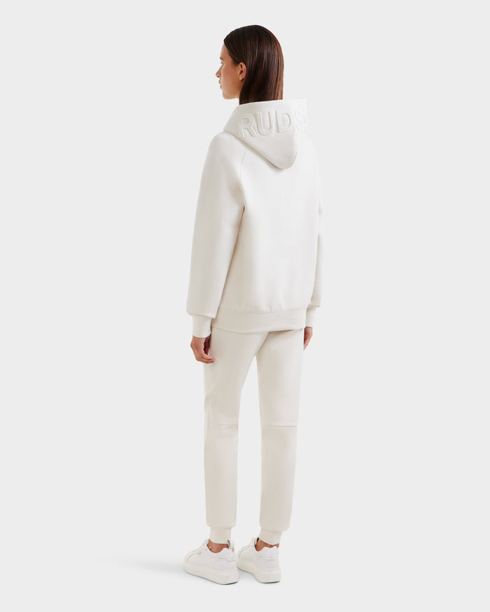 A person faces away, wearing the Rudsak BARRY - 9524011 Vanilla W unisex hoodie and sweatpants set with white sneakers. The oversized hoodie features embroidered text, hood up, against a plain light background.