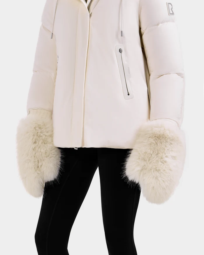 A person models the Rudsak BELLA - 8425006 Vanilla jacket, a white puffer with large cream fur cuffs, paired with black pants against a light background and showcasing chic faux fur mittens.