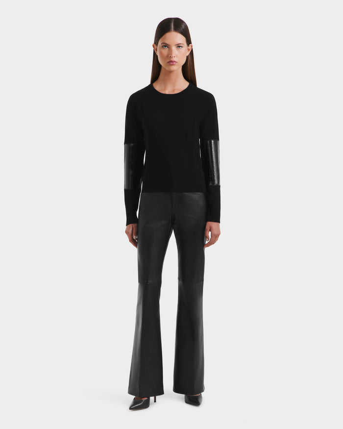 A woman stands facing forward in the Rudsak BELLEROSE - 8525554 Black crewneck sweater with shiny sleeve panels and black flared leather pants, paired with pointed black heels, against a plain white background.