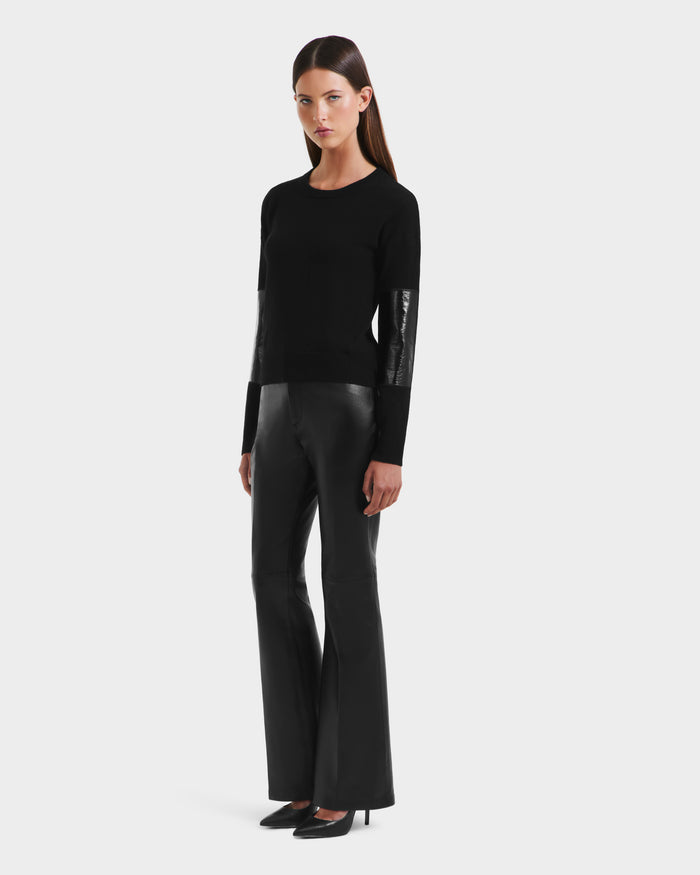A woman stands against a plain light background wearing the Rudsak BELLEROSE - 8525554 Black sweater with shiny sleeve panels, paired with black flared pants and high-heeled shoes.