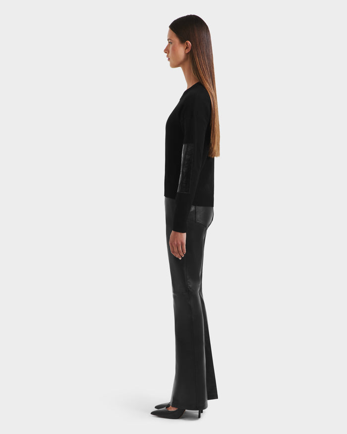A woman stands in profile against a plain background, wearing the Rudsak BELLEROSE - 8525554 Black sweater with leather elbow patches, paired with black leather pants and pointed-toe shoes. Her straight brown hair is worn down.