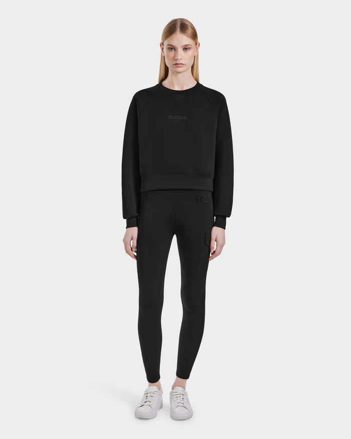 A woman with straight blonde hair wears the Rudsak BETSY - 8525087 Black crewneck sweater and black leggings, paired with white sneakers, standing against a plain light gray background.