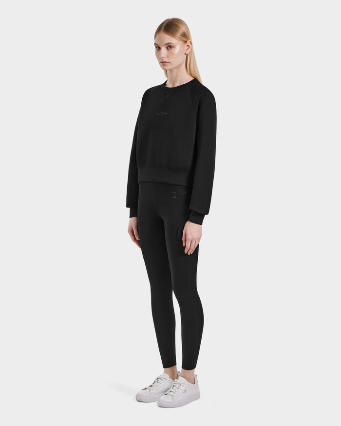 A woman with straight blonde hair and a neutral expression wears the Rudsak BETSY - 8525087 Black women’s crop sweater against a plain background, styled with matching leggings and white sneakers.