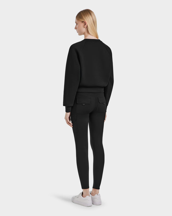 A woman with straight blonde hair faces away, wearing the Rudsak BETSY - 8525087 Black crewneck sweater with fitted black pants and white sneakers, set against a plain light gray background.