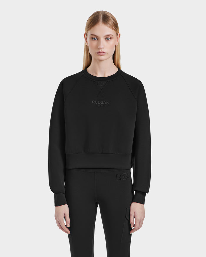 A young woman with straight blonde hair models the Rudsak BETSY - 8525087 Black sweatshirt, a modern cropped silhouette in premium cotton blend with subtle RUDSAK branding, paired with black pants against a plain white background.