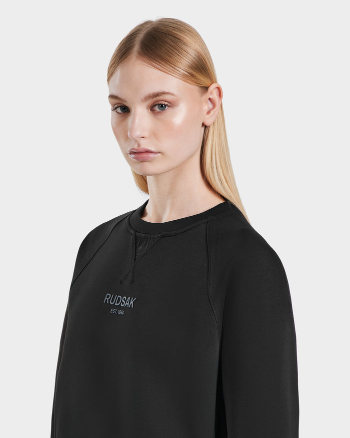 A young woman with straight blonde hair wears the Rudsak BETSY - 8525087 Black womens crop sweater, made of premium cotton blend, standing against a plain light background.