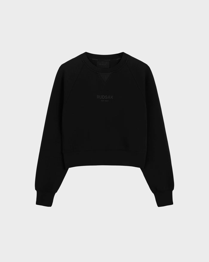 The BETSY - 8525087 Black is a black crewneck sweater by Rudsak with ribbed cuffs and hem, featuring the brand subtly printed on the front in a modern cropped silhouette, shown against a plain light grey background.