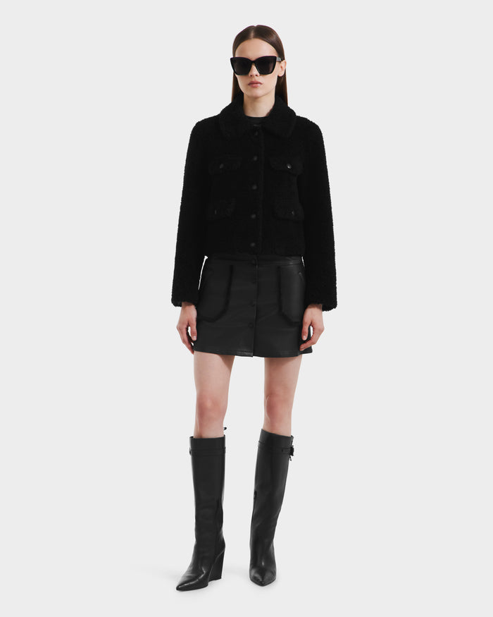 A woman faces forward against a white background, wearing the Rudsak BLAIR - 8125513 Black modern jacket, paired with a black mini skirt, tall black boots, and oversized black sunglasses.