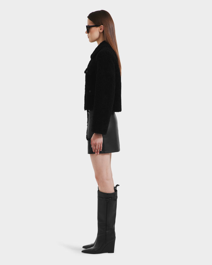 A woman stands in profile against a plain backdrop, wearing the Rudsak BLAIR - 8125513 Black cropped teddy jacket with a leather mini skirt, knee-high black boots, and sunglasses. Her straight hair is loose and her arms rest at her sides.