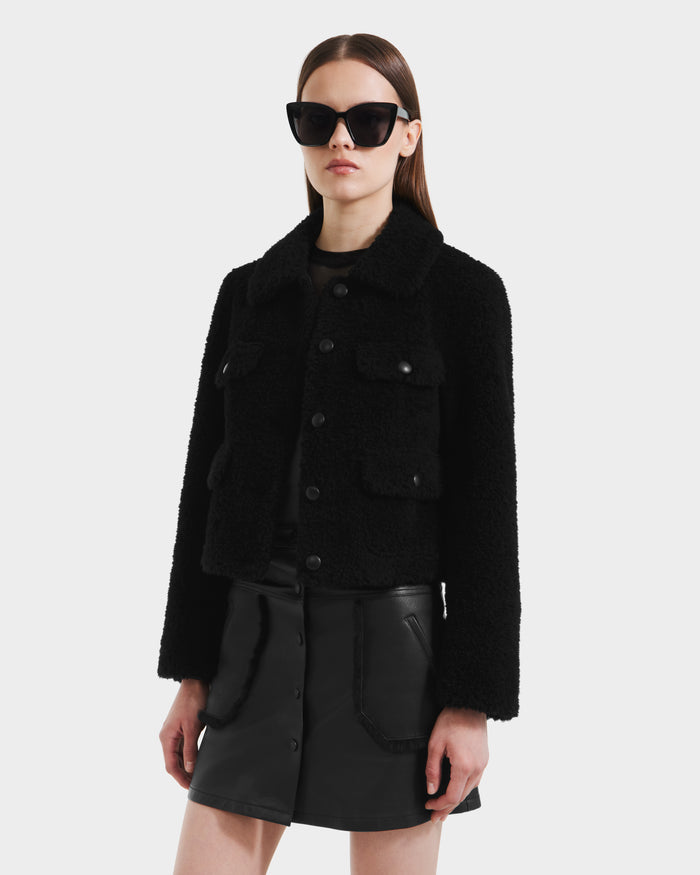 A woman wearing Rudsaks BLAIR - 8125513 Black modern cropped teddy jacket with flap pockets, large black sunglasses, and a black leather mini skirt stands against a plain light background.
