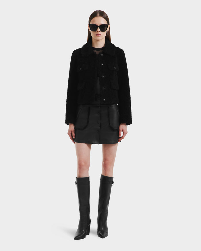 A woman faces forward against a white background wearing the Rudsak BLAIR - 8125513 Black jacket, styled with a black mini skirt, knee-high boots, large sunglasses, and straight hair parted in the middle.