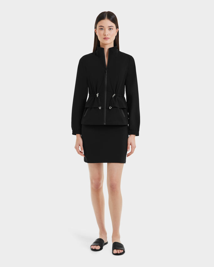 A woman stands facing forward, wearing the Rudsak BONNIE - L125012 Black lightweight windbreaker with an adjustable waistcord and a matching black skirt. She has straight dark hair and wears black slide sandals against a plain white background.