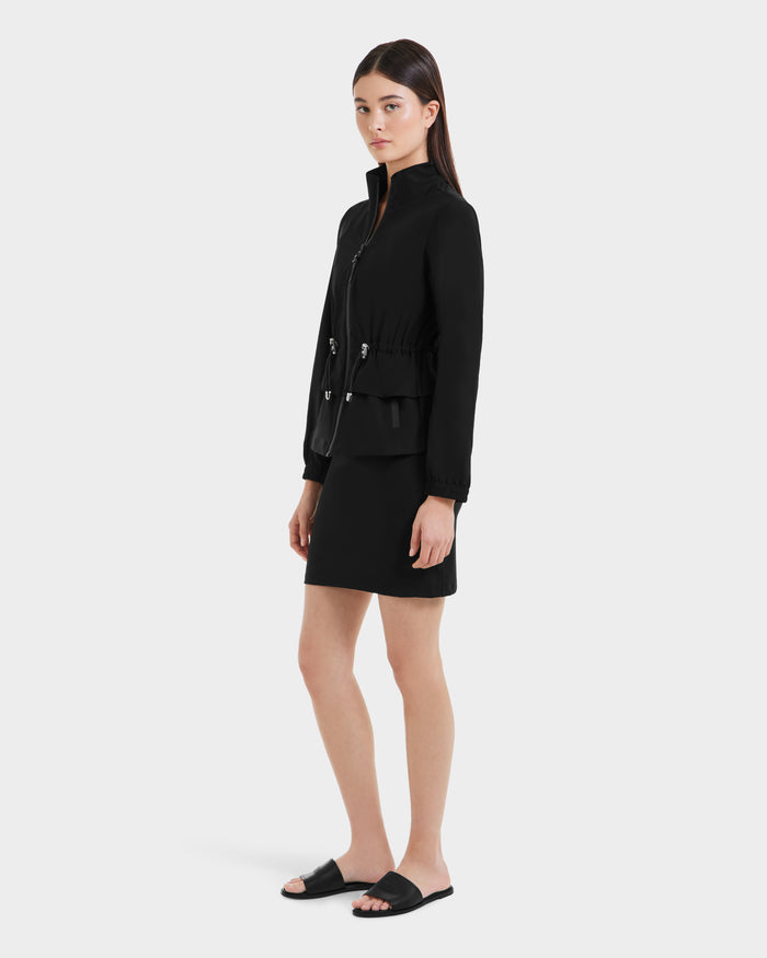 A young woman with straight brown hair wears Rudsak’s BONNIE - L125012 Black lightweight jacket with an adjustable waistcord, paired with a black skirt and black slides, standing against a plain light background.