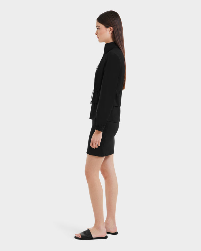 A woman with straight brown hair wears the Rudsak BONNIE - L125012 Black lightweight jacket, paired with a black skirt and black slide sandals, standing in profile against a plain white background.