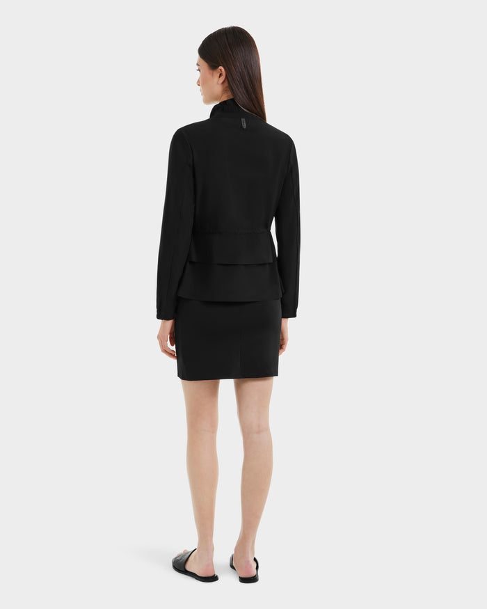 A woman with straight brown hair stands facing away against a white background, wearing the Rudsak BONNIE - L125012 Black lightweight jacket with an adjustable waistcord, a black skirt, and black slide sandals.