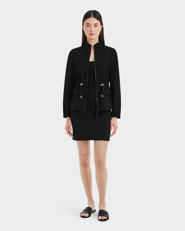 A woman with straight brown hair and a neutral expression models the Rudsak BONNIE - L125012 Black lightweight jacket over a black dress and slide sandals, standing against a plain light gray background.