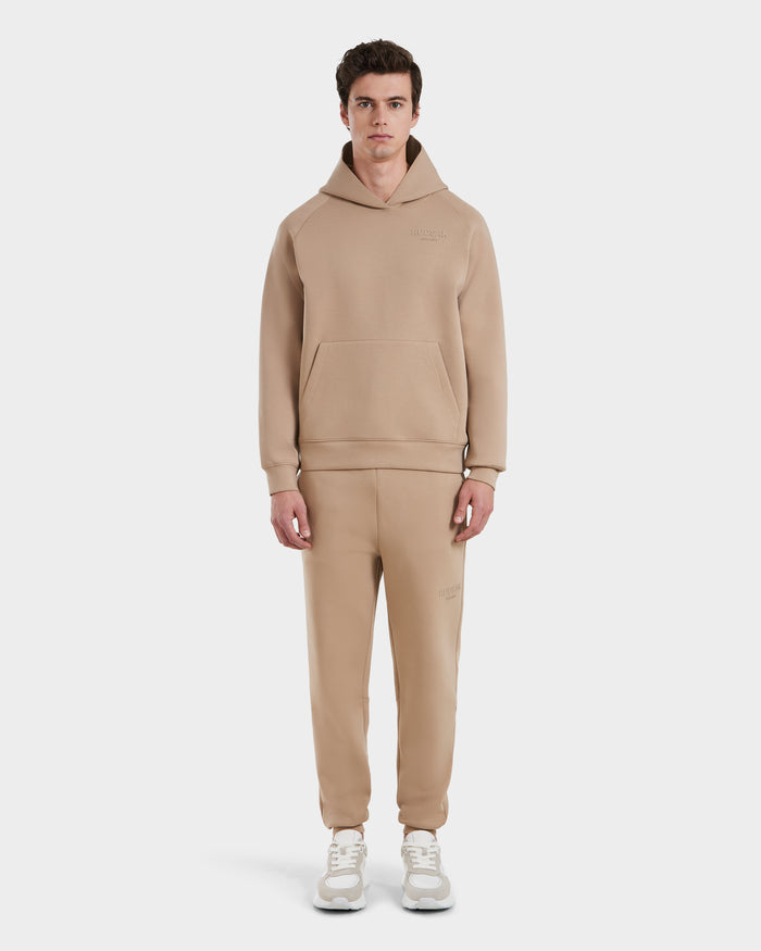 A man stands facing forward in the Rudsak BRIAR - 9525001 Almond M unisex hoodie and sweatpants set, made from a premium cotton blend, paired with white sneakers against a plain white background.
