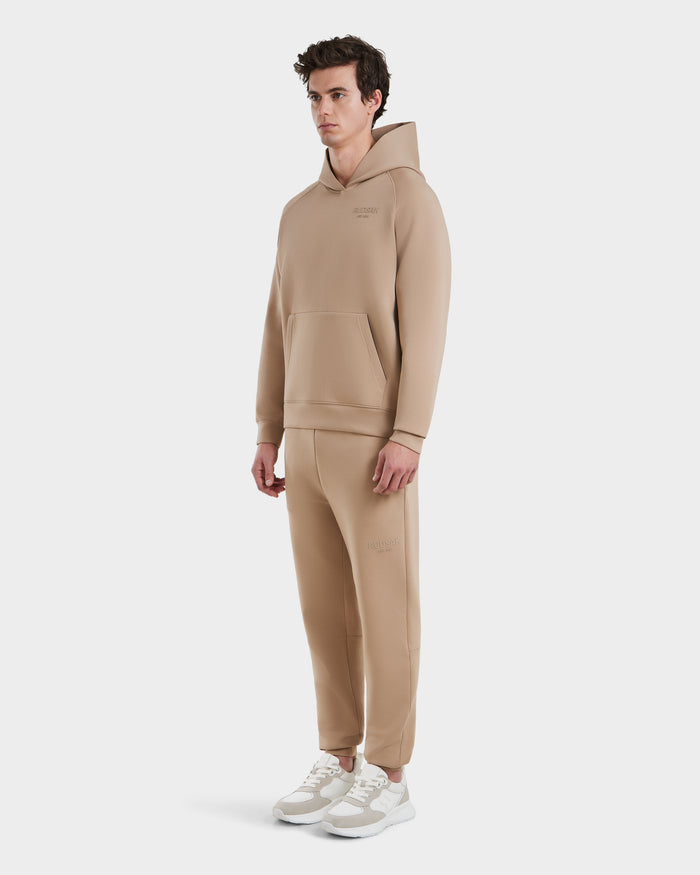 A man faces forward in a Rudsak BRIAR - 9525001 Almond M unisex hoodie and joggers set, made from premium cotton blend, with white sneakers against a plain white background.