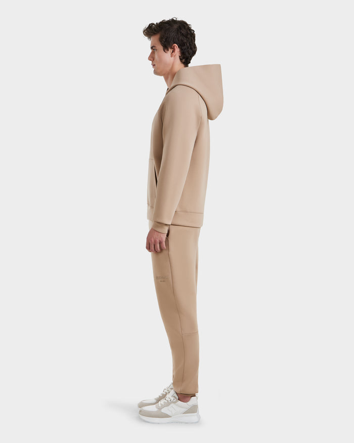 A man in profile faces left, wearing the Rudsak BRIAR - 9525001 Almond M unisex hoodie and matching cotton-blend sweatpants with white sneakers, stands against a plain white background.
