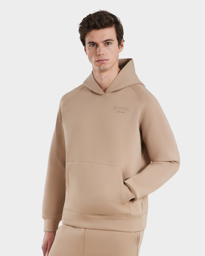 A person stands facing forward with one hand in the pocket, showcasing the Rudsak BRIAR - 9525001 Almond M unisex hoodie and matching pants for a casual look against a plain white background.