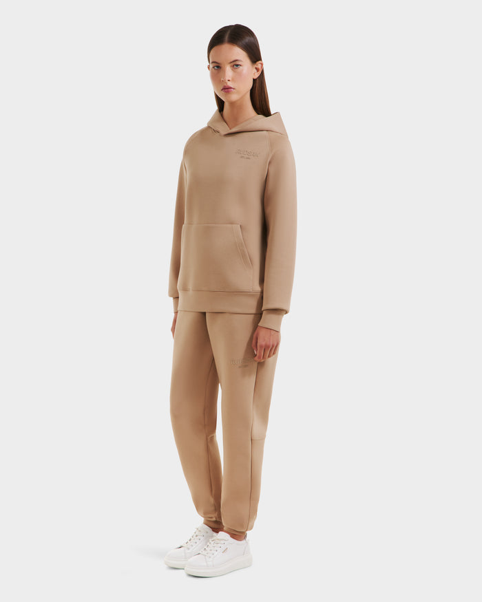A woman faces forward wearing the Rudsak BRIAR - 9525001 Almond W premium cotton blend unisex hoodie and matching beige sweatpants set with white sneakers, standing against a plain white background.