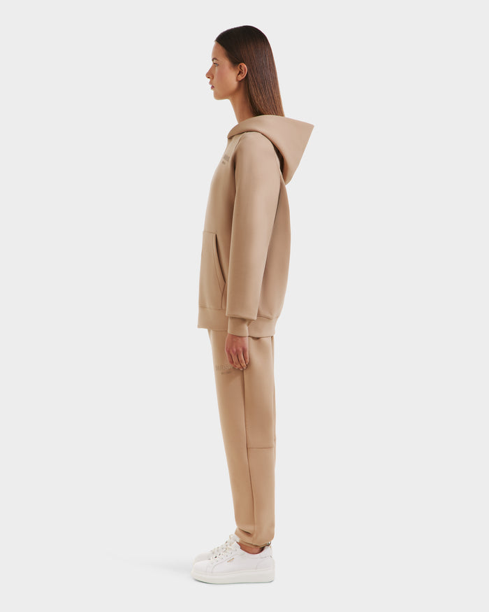 A woman in profile models the Rudsak BRIAR - 9525001 Almond W hoodie and sweatpants set, paired with white sneakers for a relaxed, casual look against a white background.