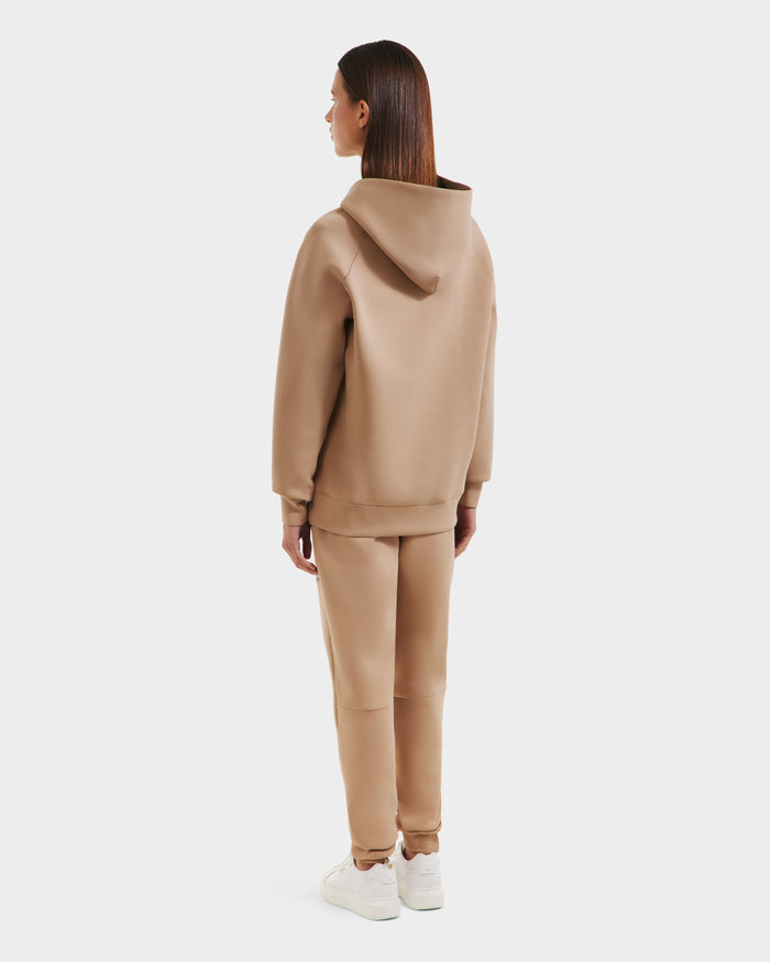 A woman with straight brown hair wears the Rudsak BRIAR - 9525001 Almond W hoodie and matching sweatpants, paired with white sneakers. She faces away against a plain light background, showcasing a relaxed, premium cotton blend casual look.