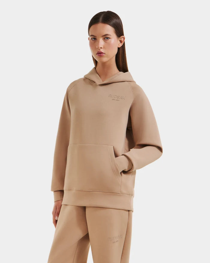 A woman wearing the Rudsak BRIAR - 9525001 Almond W premium cotton hoodie and matching sweatpants stands against a plain white background with one hand in her front pocket.