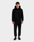 A man faces forward in a Rudsak BRIAR - 9525001 Black M unisex premium cotton blend hoodie with RUDSAK on the chest, black pants, and black sneakers, set against a plain light gray background.