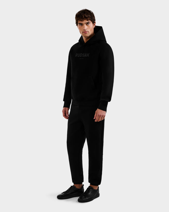 A young man faces forward wearing the Rudsak BRIAR - 9525001 Black M hoodie and matching black joggers, paired with black sneakers. The look is monochrome and casual, set against a plain light gray background.