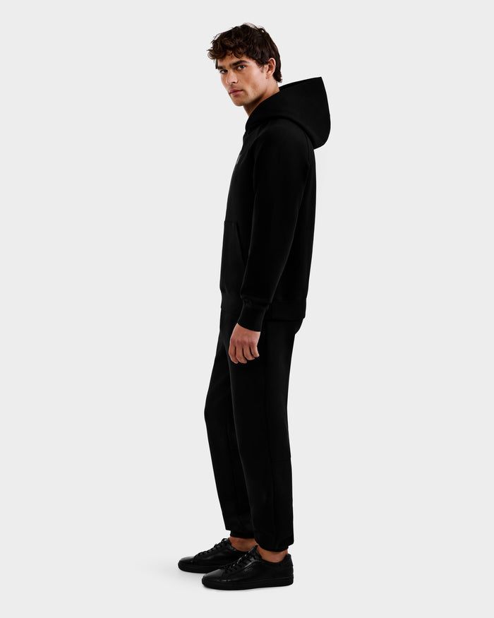 A man stands sideways against a white background, wearing the Rudsak BRIAR - 9525001 Black M hoodie with black pants and sneakers. He faces the camera with a neutral expression.