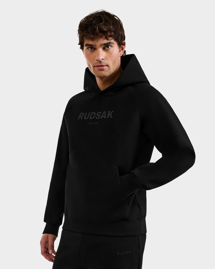 A man stands confidently against a light gray background, wearing the Rudsak BRIAR - 9525001 Black M hoodie and matching black pants, with one hand in his pocket and looking at the camera.