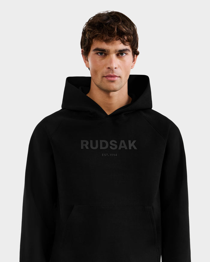 A young man with short brown hair wears the Rudsak BRIAR - 9525001 Black M unisex hoodie, made from a premium cotton blend with "RUDSAK EST. 1994" on the front, standing against a plain light grey background.