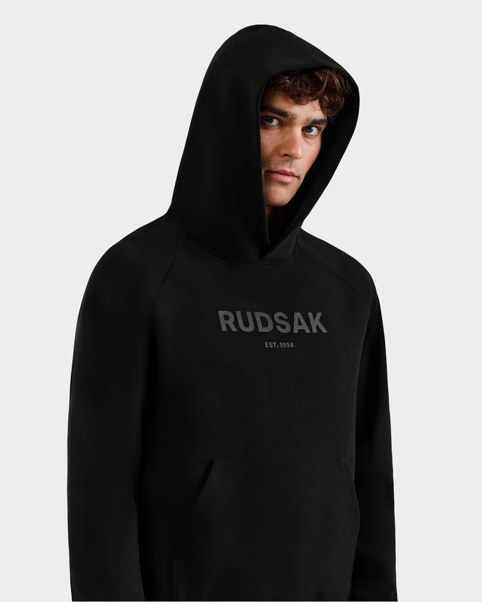 A young man stands against a light gray background wearing the Rudsak BRIAR - 9525001 Black M unisex hoodie, crafted from a premium cotton blend, with the hood up and "RUDSAK EST. 1994" printed in dark letters on the front.