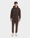 A man stands facing forward in Rudsaks BRIAR - 9525001 Espresso M unisex hoodie and matching joggers with white sneakers, creating a casual look against a plain white background.