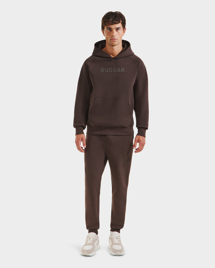 A man stands facing forward in Rudsaks BRIAR - 9525001 Espresso M unisex hoodie and matching joggers with white sneakers, creating a casual look against a plain white background.