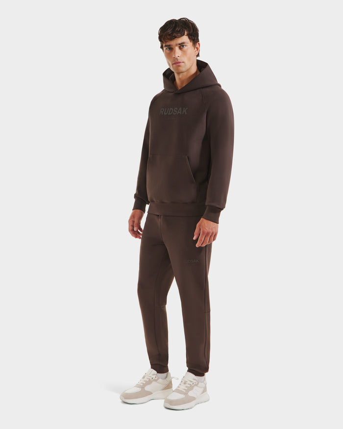 A man stands against a plain white background in Rudsak’s BRIAR - 9525001 Espresso M hoodie and matching joggers, paired with white sneakers for a relaxed, casual look.